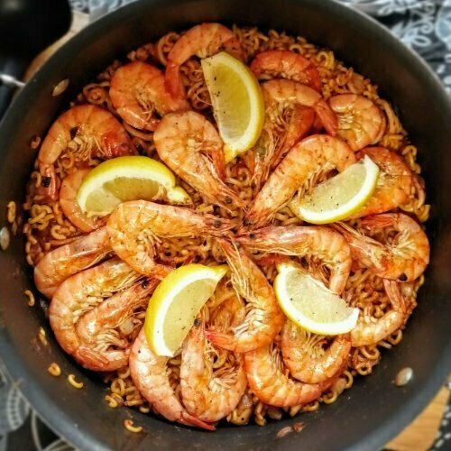 Fideua in a large black pot and topped with large prawns and wedges of lemon