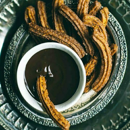 A silver platter holds a stack of churros, lightly dusted with icing. sugar, and set beside a small pot of warm, molten chocolate.