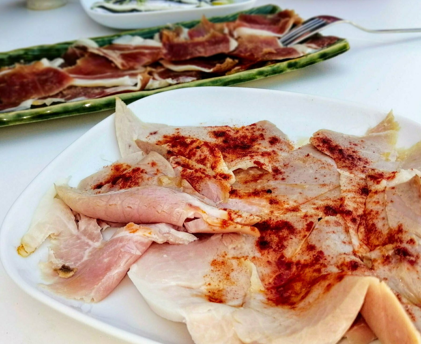 A plate of lcaon a la gallega sits on a table with a small plate of jamon iberico in the background