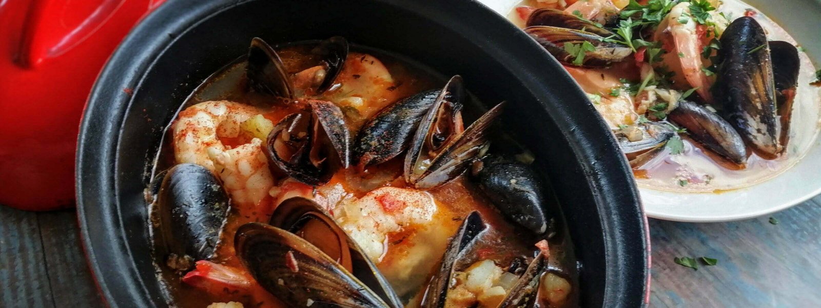 A steaming pot of Spanish seafood Stew sits beside a white bowl full of stew containing mussels, prawns and fish.