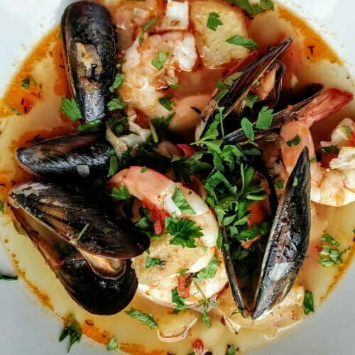 A large white bowl of Spanish seafood stew sits on a table with steaming prawns, fish, and mussels, garnished with a little chopped parsley.