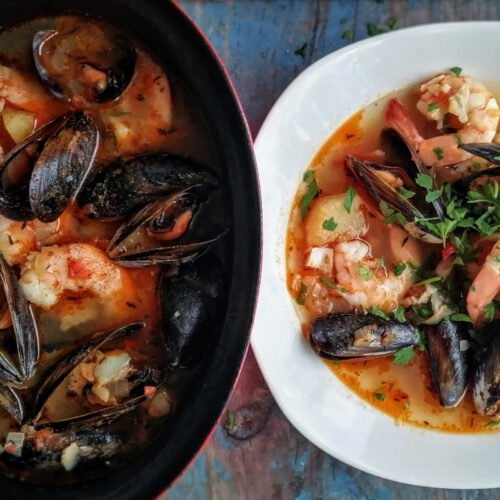 Spanish seafood stew sits in a alrge pot beside white bowl filled with stew.