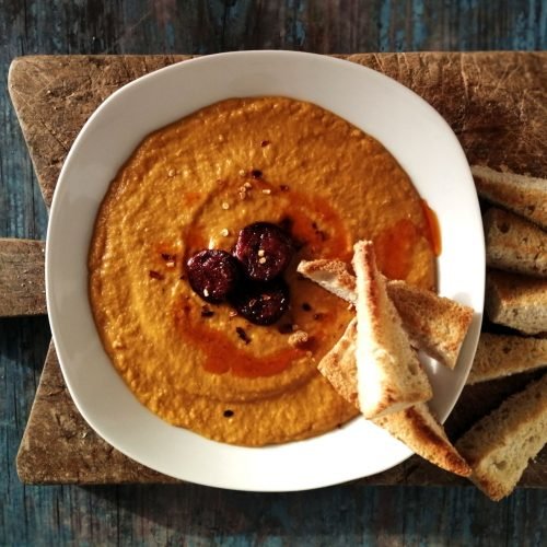 A bowl of pumpkin soup sits on a rustic chopping board and is topped with chorizo and some fresh bread