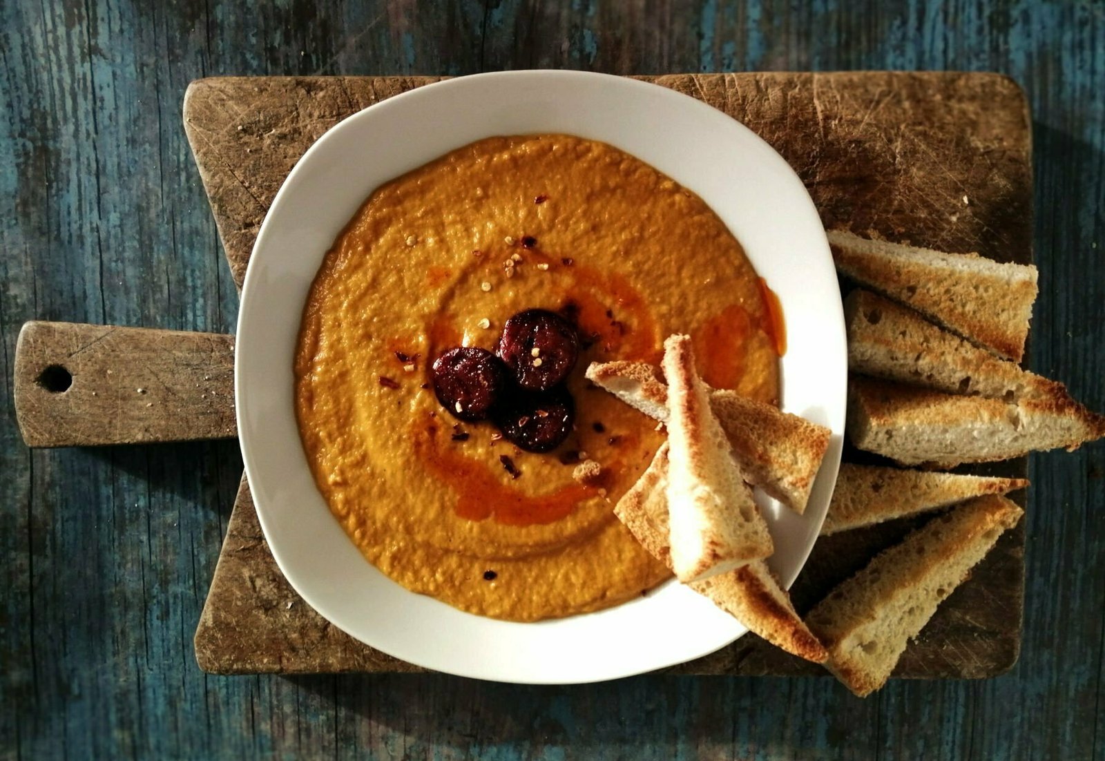 A bowl of pumpkin soup sits on a rustic chopping board and is topped with chorizo and some fresh bread