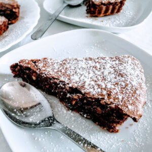 a plate of chocolate hazelnut tart sits beside a silver spoon on a white tablecloth