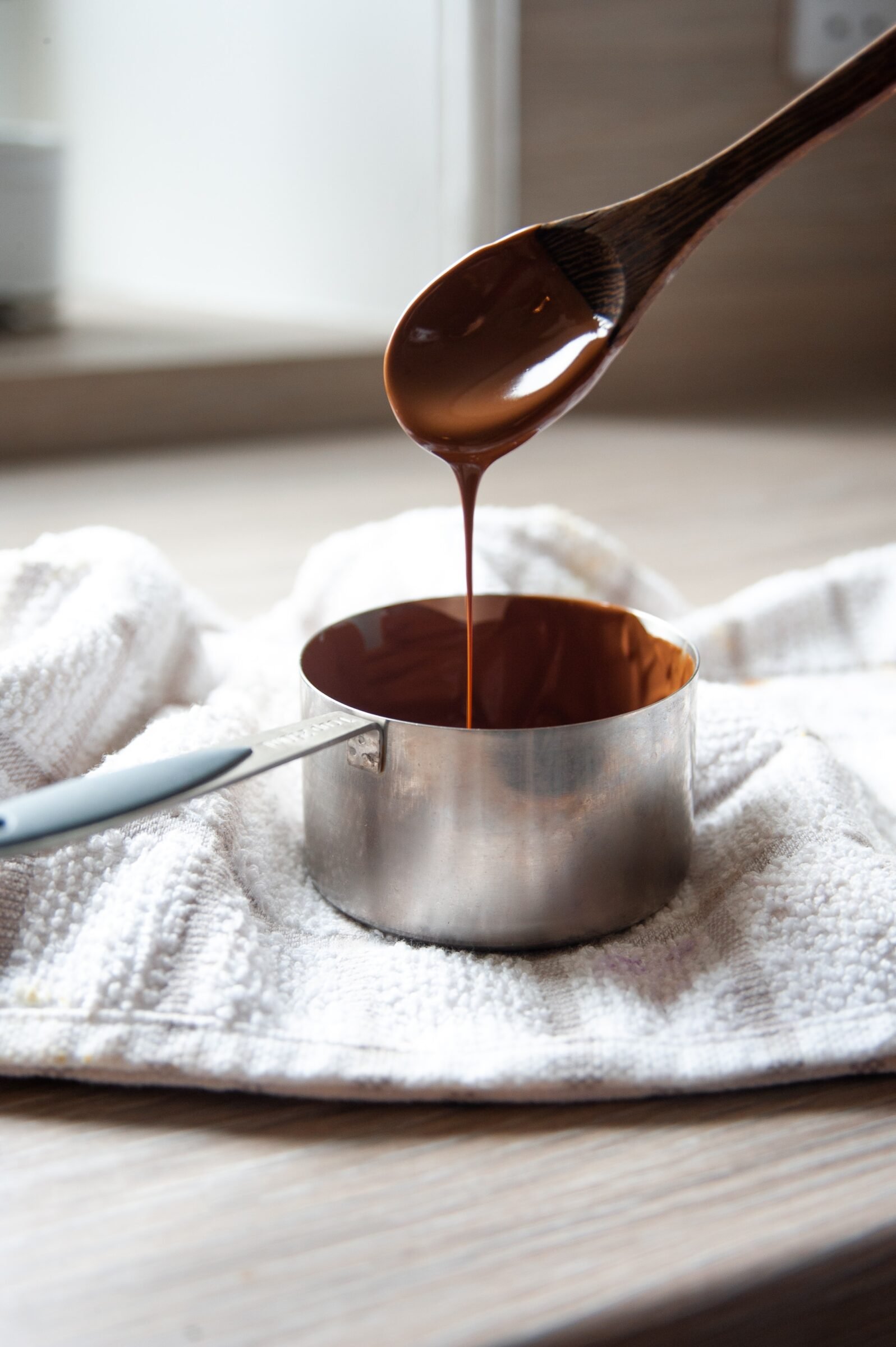 a saucepan of melted chocolate sits on a white towel. molten chocolate drizzles from a wooden spoon into the pan.