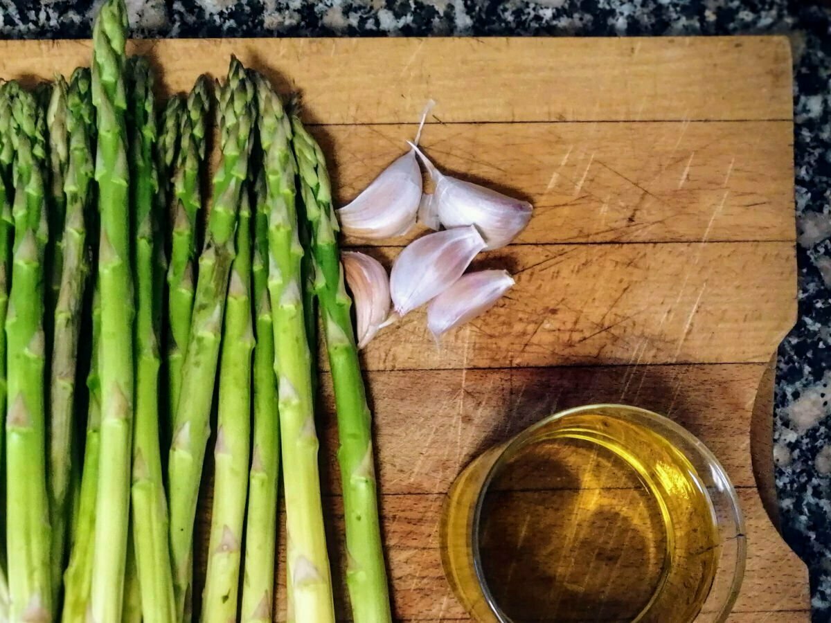 A rustic chopping board sits with some asparagus spears, garlic, and extra virgin olive oil