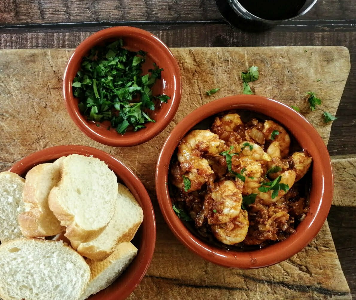 A clay pot of gambas al ajillo sits on a rustic chopping board alongside some fresh sliced bread and a small dish of parsley