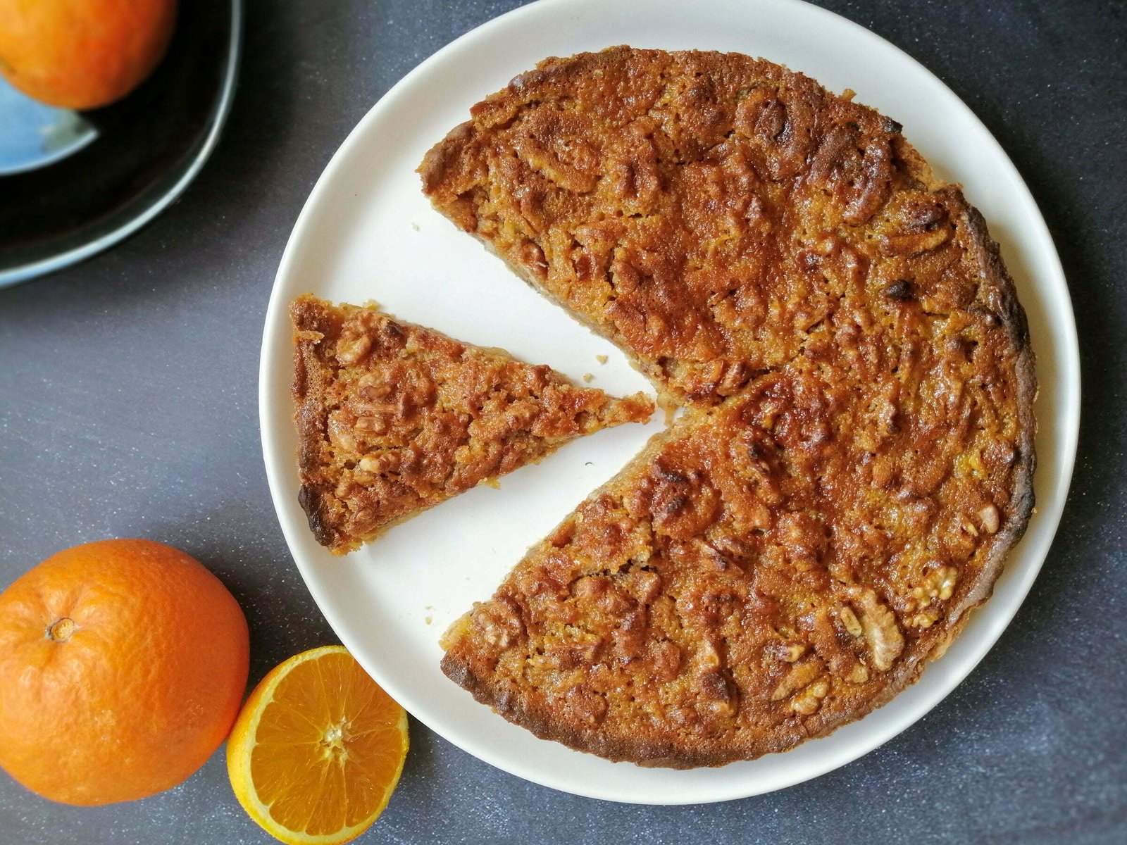 A large Walnut tart with orange and honey-infused salted caramel sits on a white plate with some oranges placed nearby