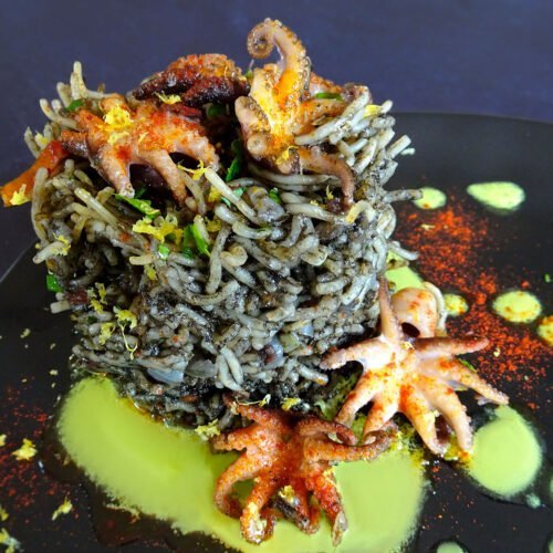 A tower of fideua negra sits on a black plate with some baby squid sitting on top