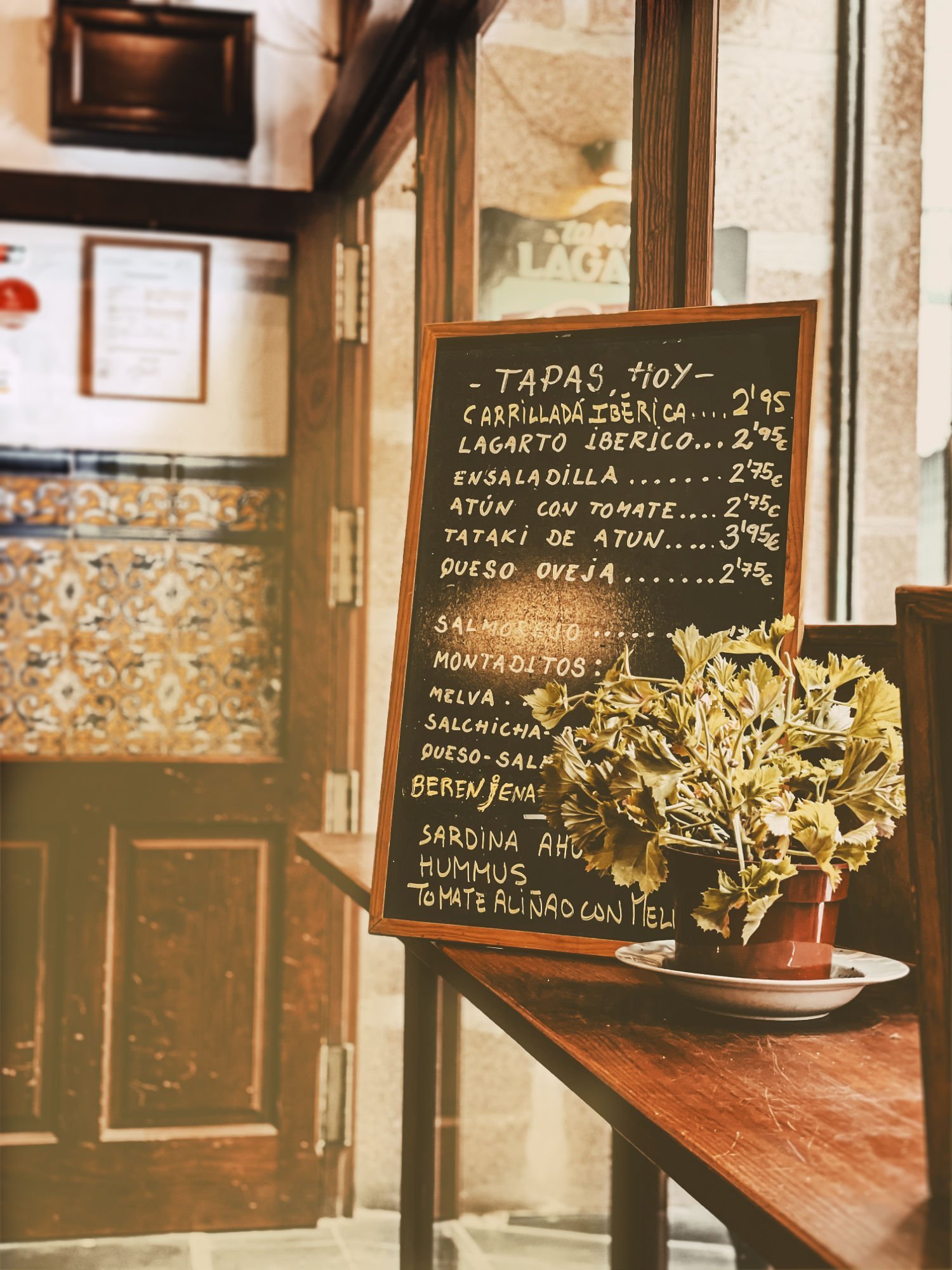An interior of a tapas bar sits bathed in golden light from outside. A chalkboard sits displaying the tapas specials of the day.
