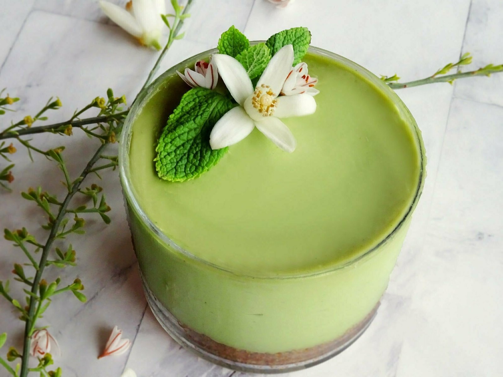 A small avocado and lime cheesecake sits in a short glass that's garnished with a few sprigs of fresh mint and an orange blossom