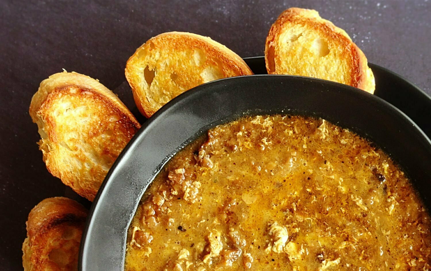 garlic soup sits in a black bowl beside some toasted bread. 
