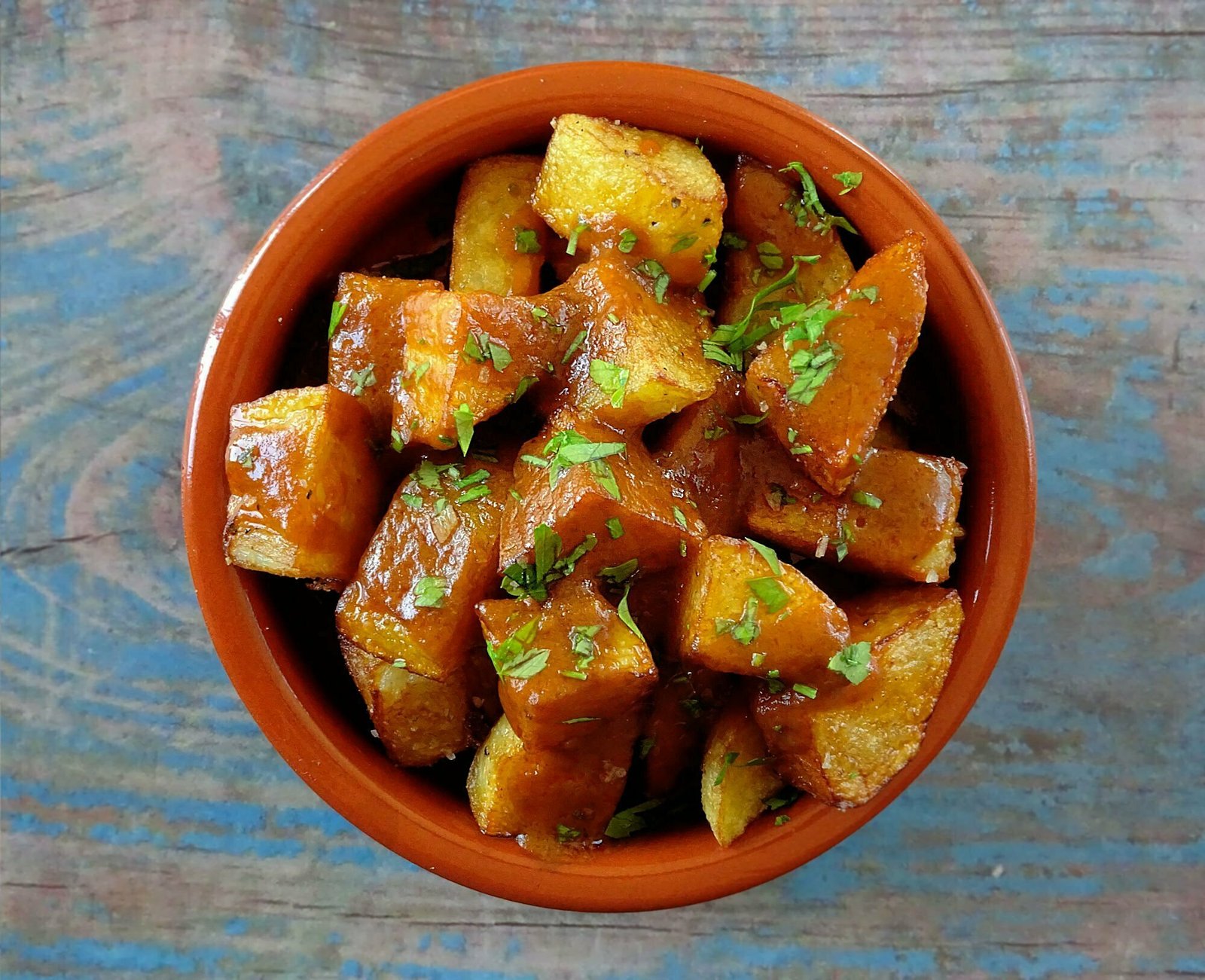 an earthenware dish sits on a blue wooden table and is filled with golden friend potato pieces smothered with Spanish bravas sauce