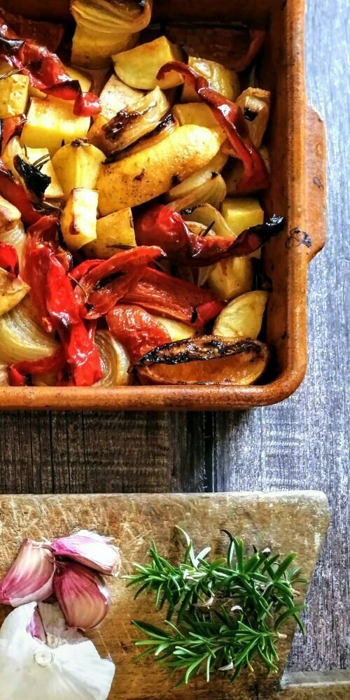 A tray of roasted potatoes sits beside a bunch of garlic cloves and some sprigs of rosemary