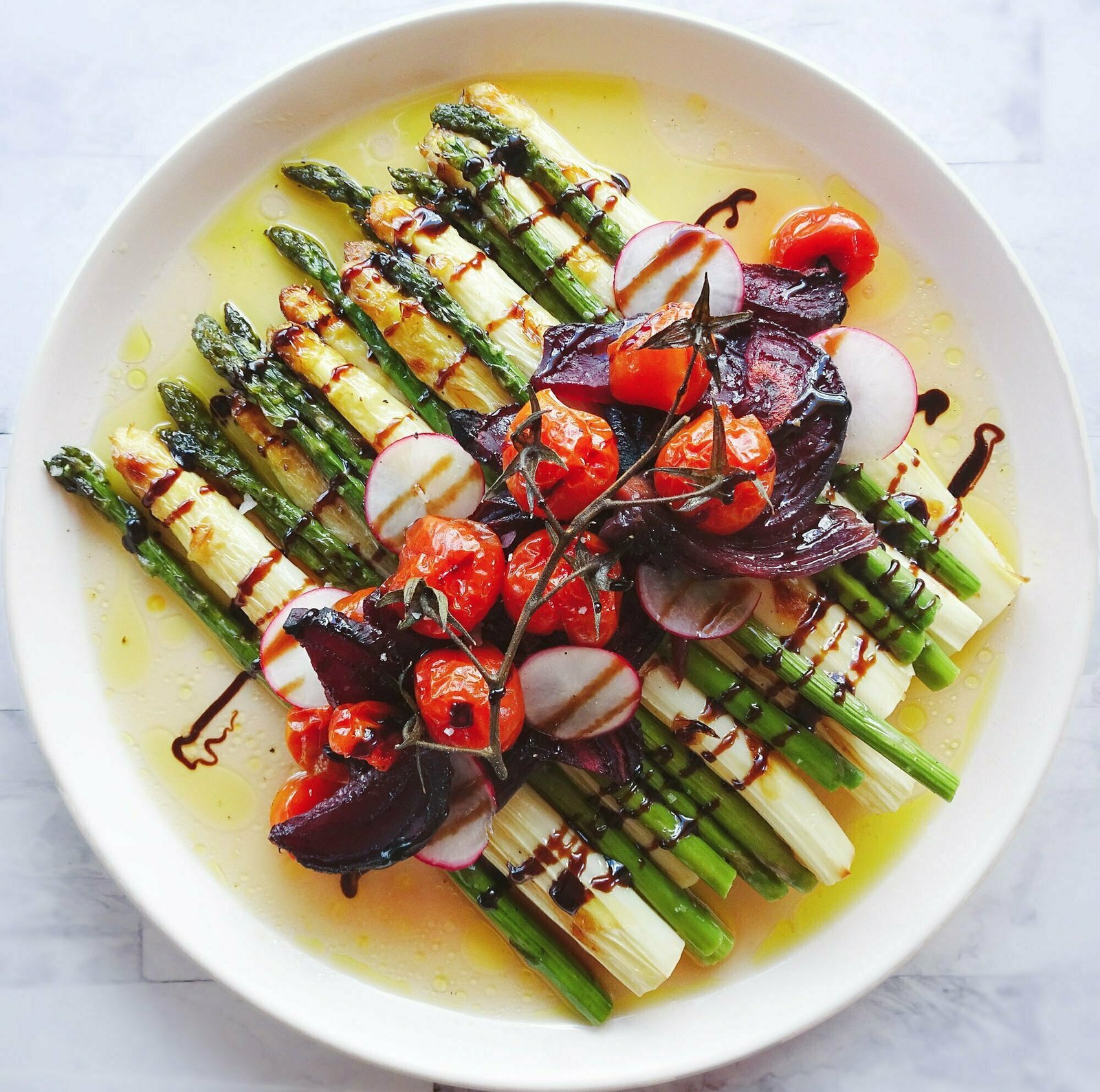 A stack of greenand white asparagus sits on a large white plate with cherry tomatoes, roasted beets and radishes for a garnish