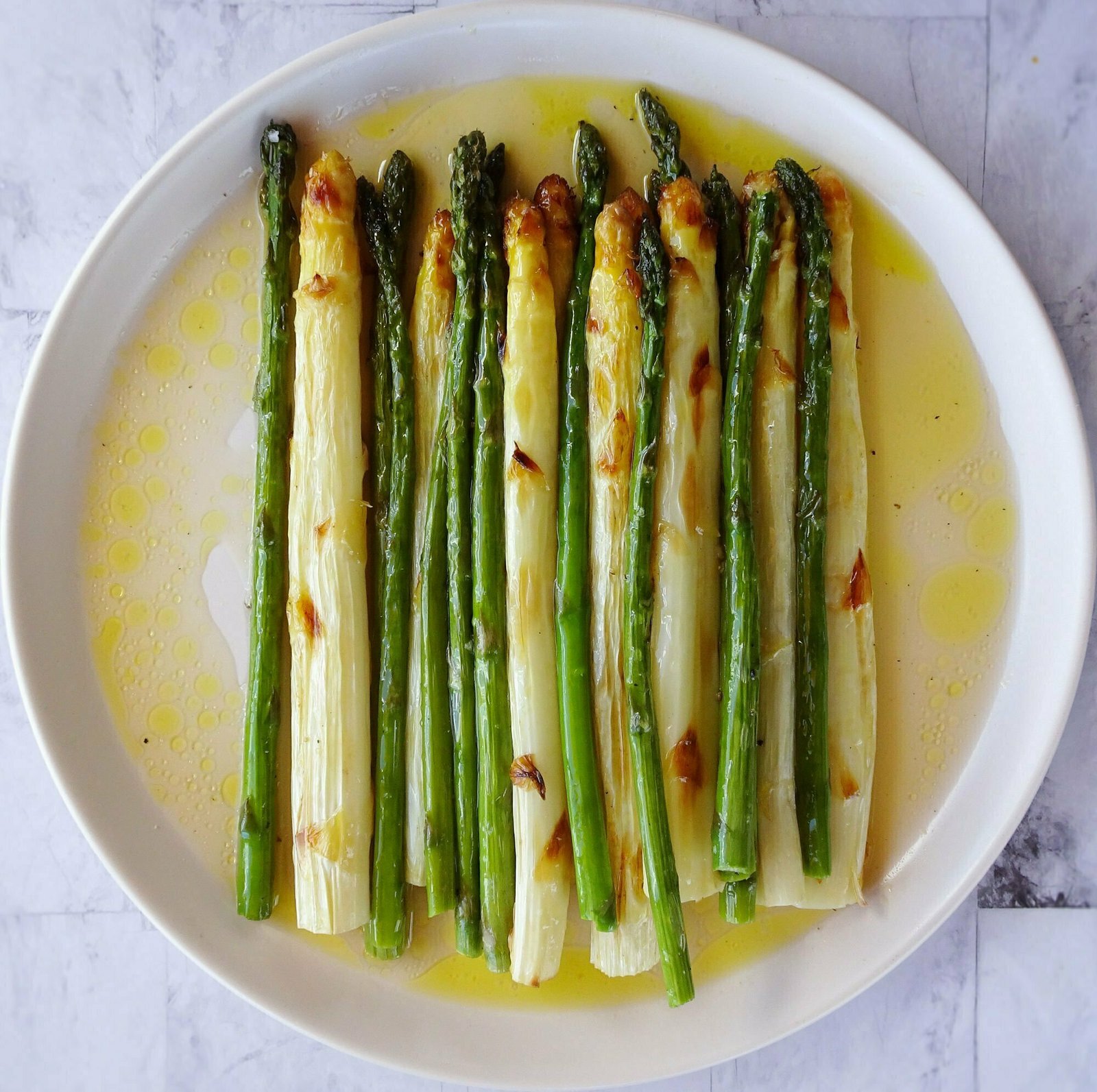 a large white plate sits on a marble counter stacked with roasted stems of white and green asparagus. A pool of oil and white wine vinegar dissolves around the stack of roasted asparagus.