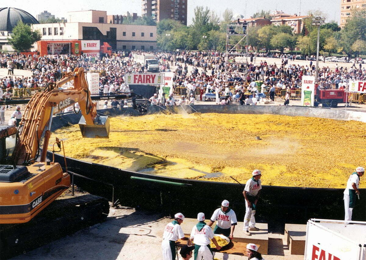 A guinness world record breaking paella is assembed in a large plaza in Vlaencia. An Earthworks machine is stirring the paella with many chefs dotted around the scene