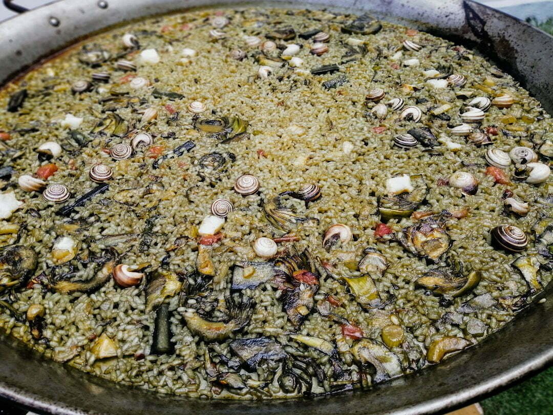 A large paell pan sits cooking with some snails, artichokes and other vegetables on top of the paella rice. 