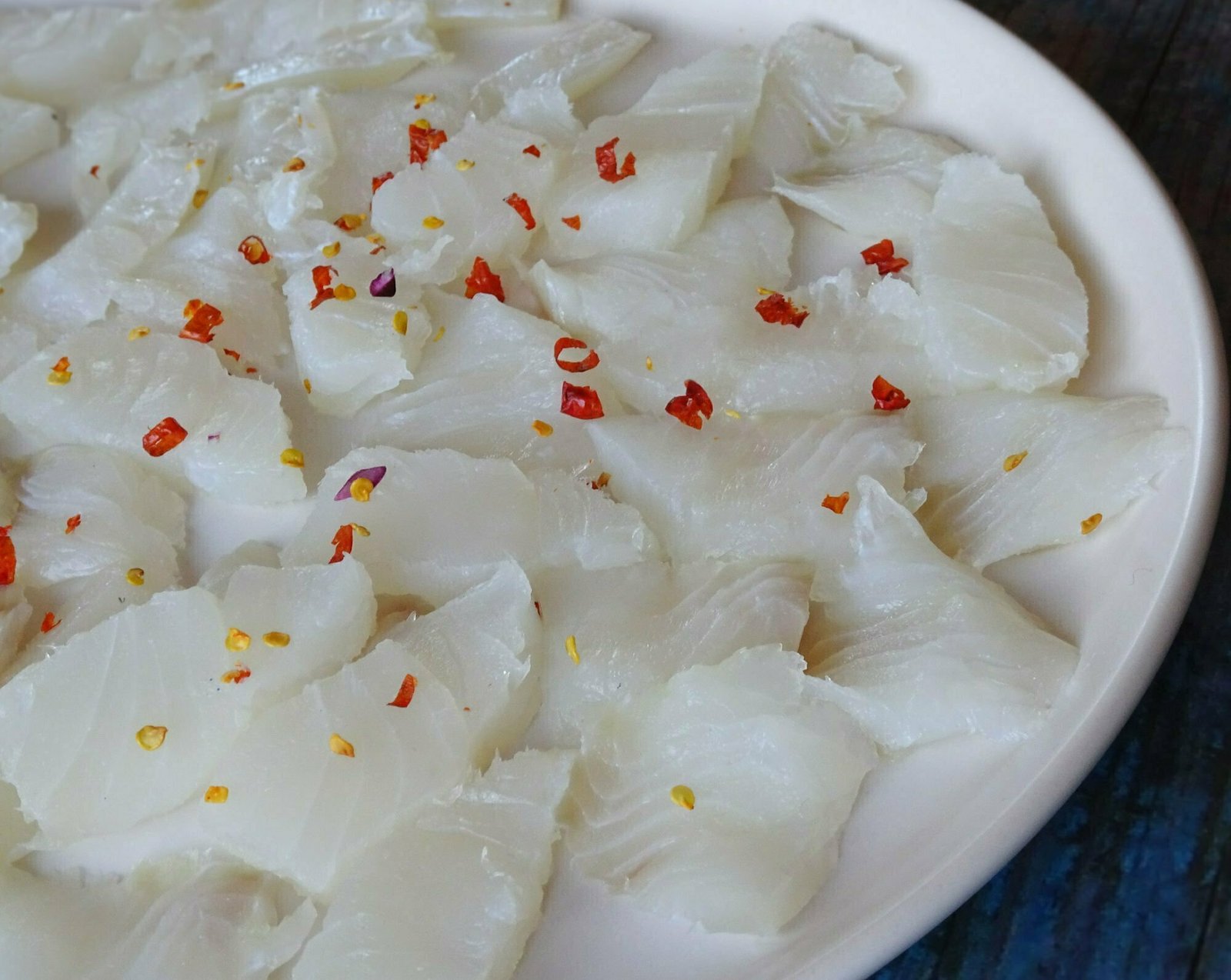 A large white plate sits on a black and blue background with fillets of white fish sprinkled with some sliced red chili 