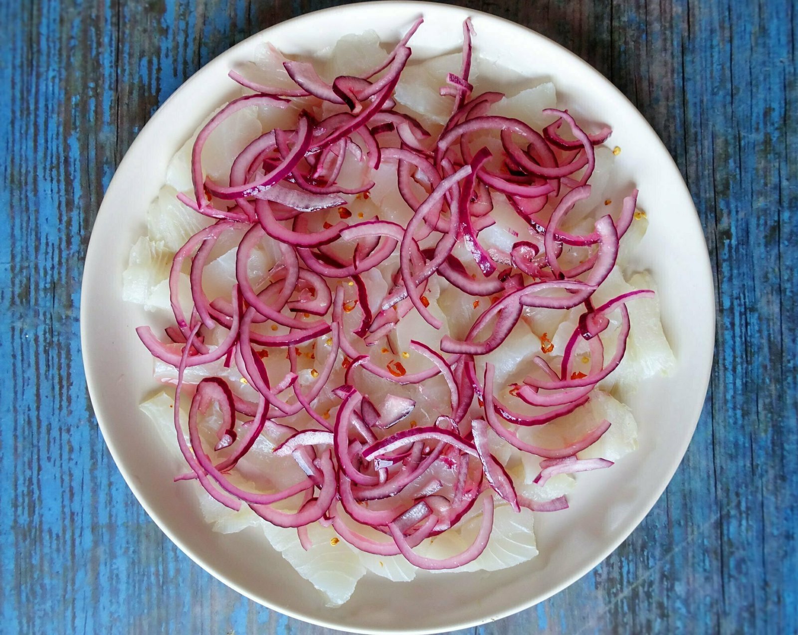 a large white plate sits o n a blue wooden background thats filled with ceviche and topped with some red chili slices and sliced red onion