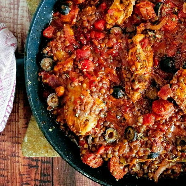 A large black pan sits on a rustic wooden board and is filled with chicken and chorizo stew.