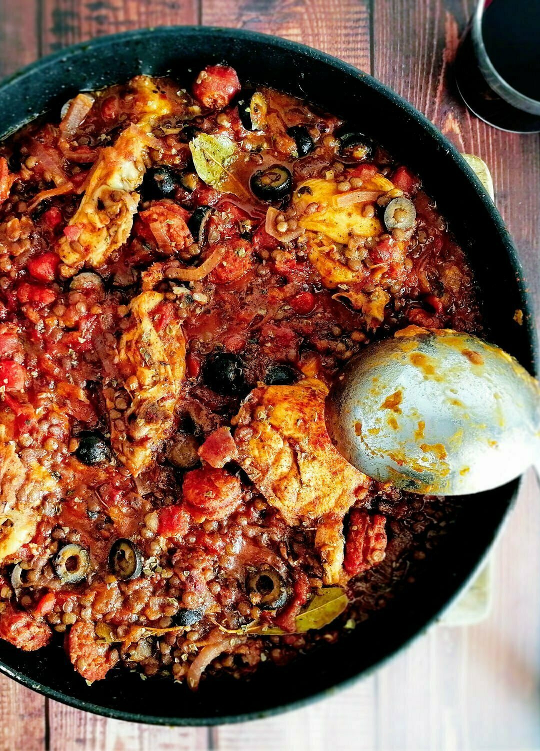 A large black pan sits on a rustic wooden board and is filled with chicken and chorizo stew.