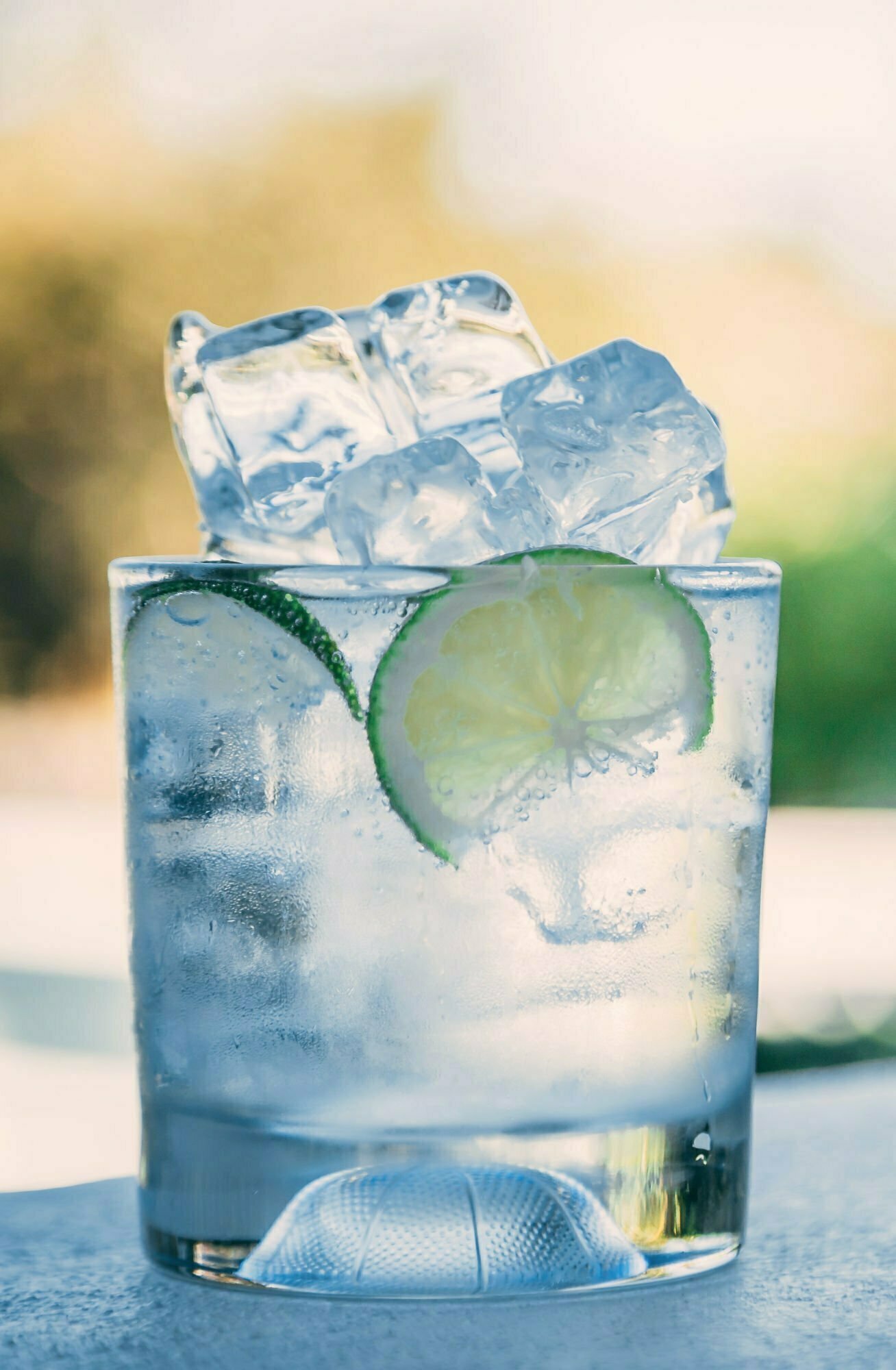 a tumber glass sits on a counter with lots of ice stacked high. A wedge of lime sits floating in a gin and tonic coktail.