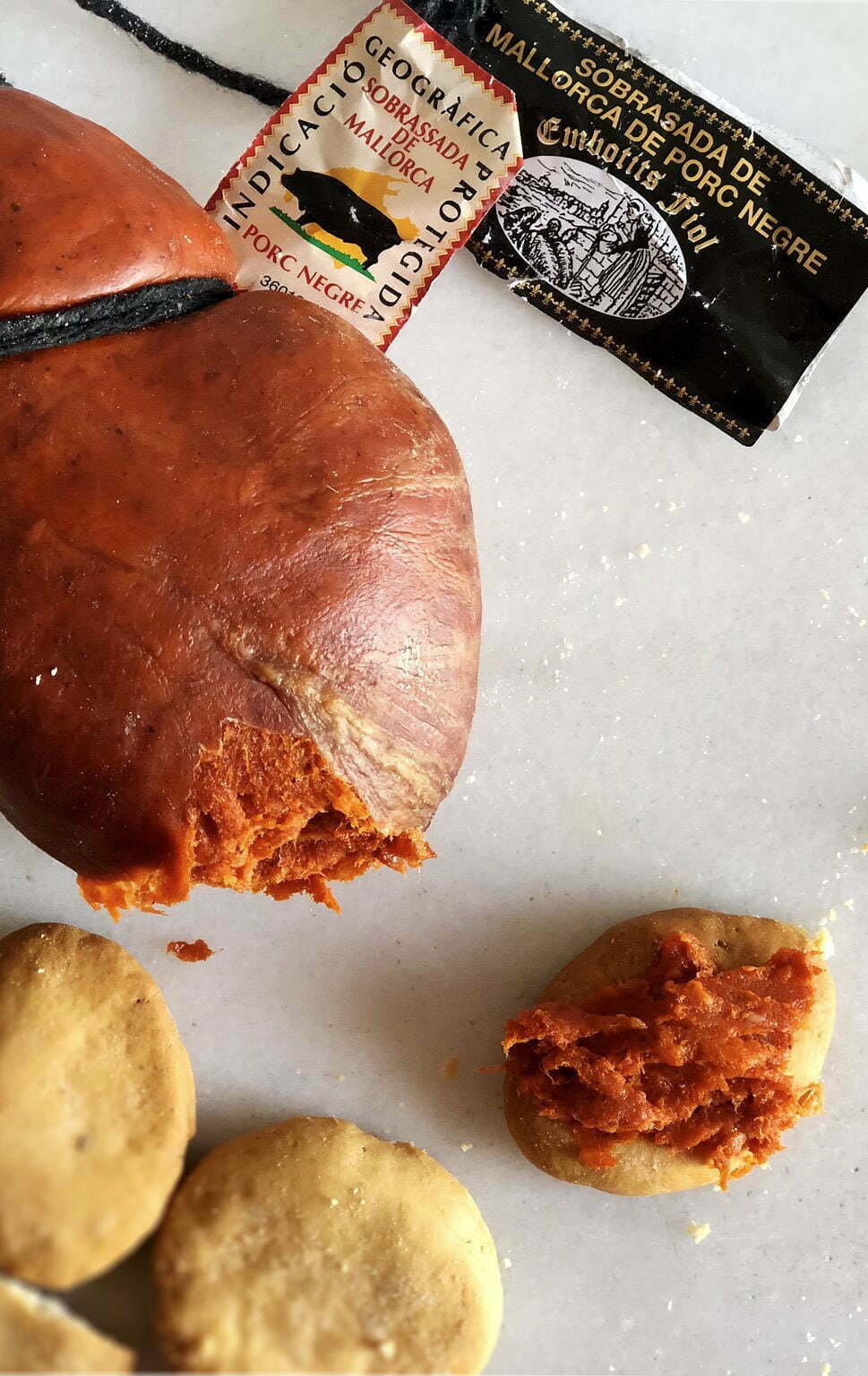 a fat sausage of sobrasada sits on a white counter with a few small crackers scattered nearby