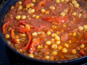 A tomato based sauce simmers on a stovetop with some chickpeas and sliced red pepper