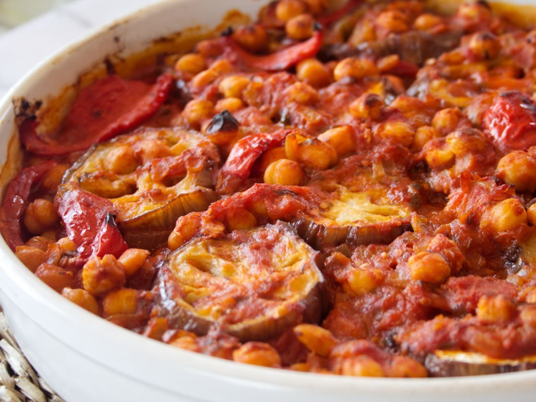A large white casserole dish sits on a white marble counter with a rich red Mediterranean Eggplant casserole with chickpeas and tomato