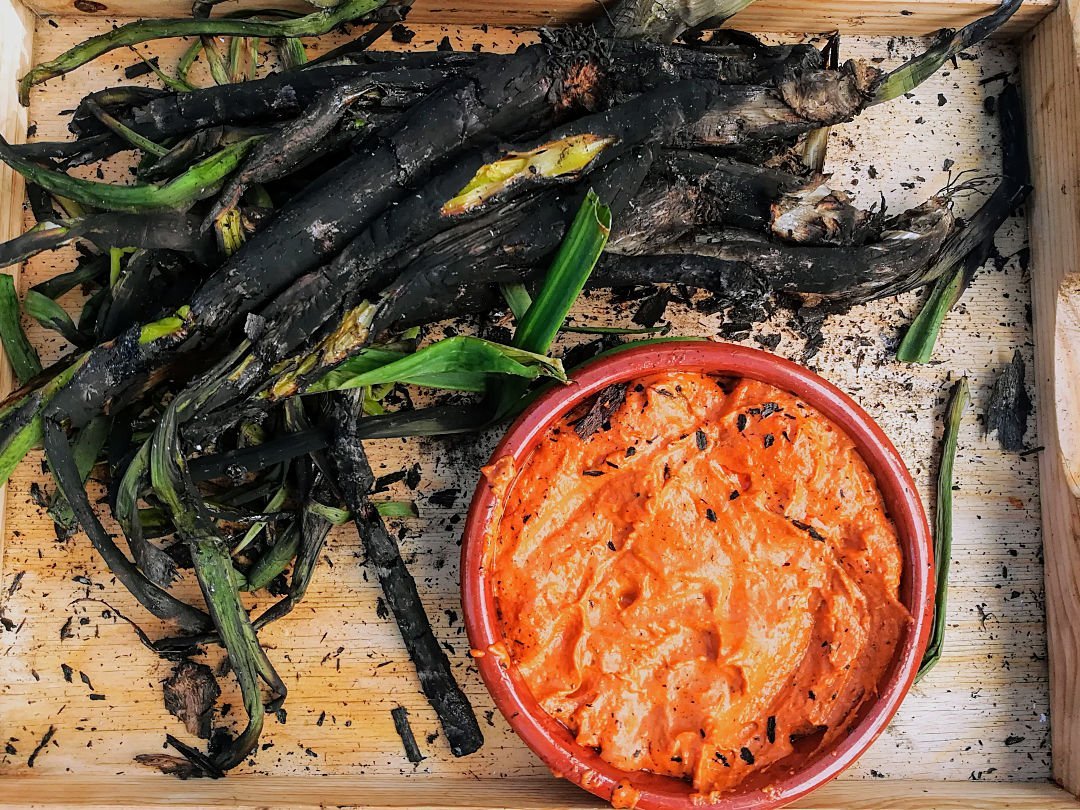 Barbercued calcots sit on a wooden servig tray with a small bowl of romesco sauce. 