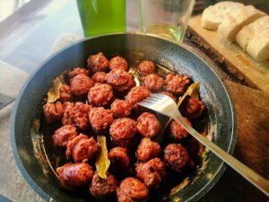 a frying pan of chorizo cooked in cider with a few bay leaves sits beside a bottle and glass of cider