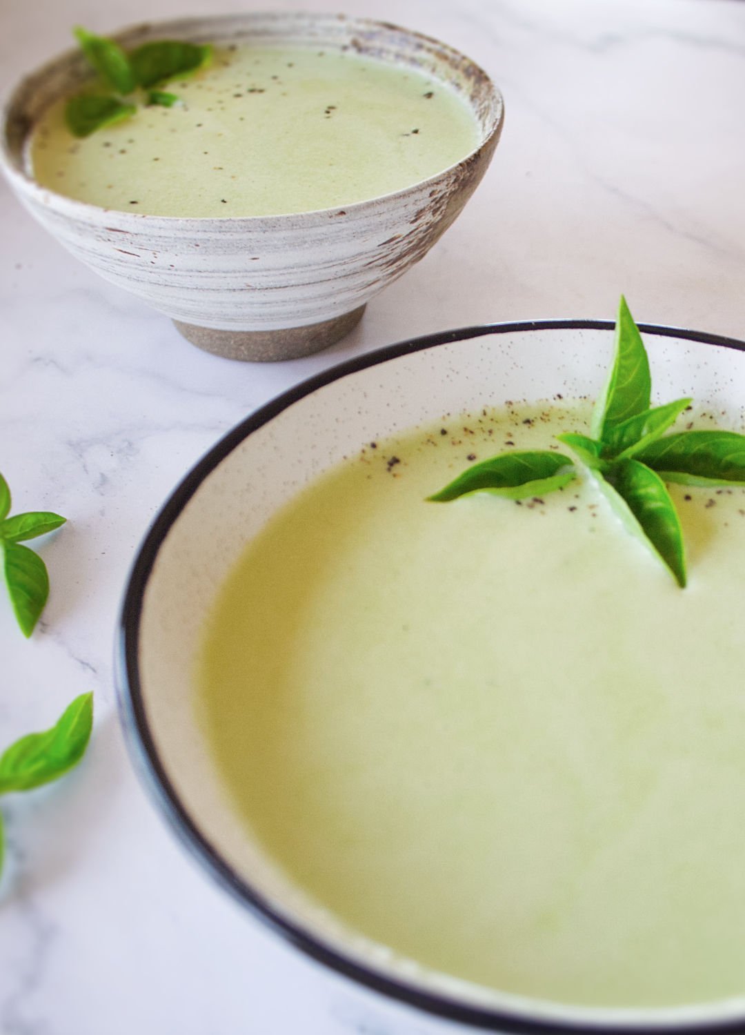 2 bowls sit filled with cold cucumber soup and garnished with some fresh basil