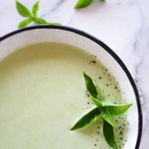 A white bowl sits on a couter with cold cucumber soup and garnished with fresh sprigs of basil
