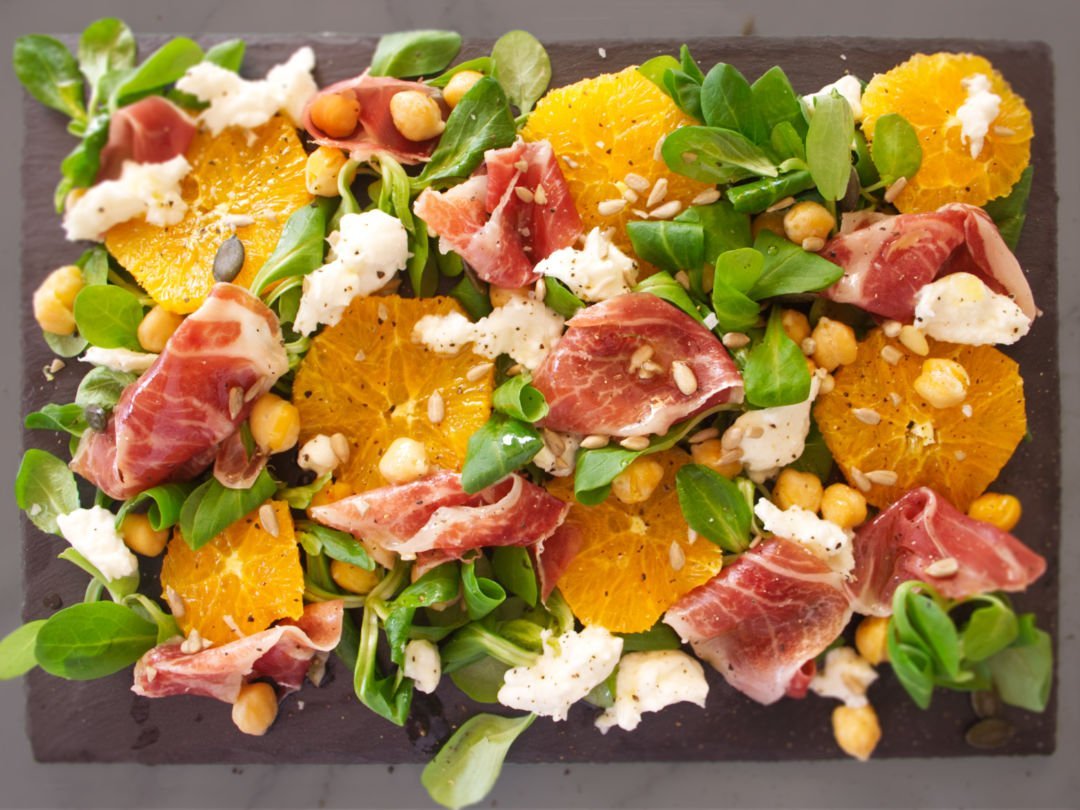 A large platter of jamon iberico sald sits topped with mozarella, salad greens, chickpeas, and seasoned with salt and pepper