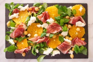 some jamon iberico is added to slate plate, with the greens, orange slices and cchickpeas.