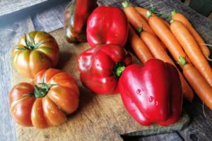 large tomatoes, red peppers, and carrots sit on a chopping board waiting to be prepared