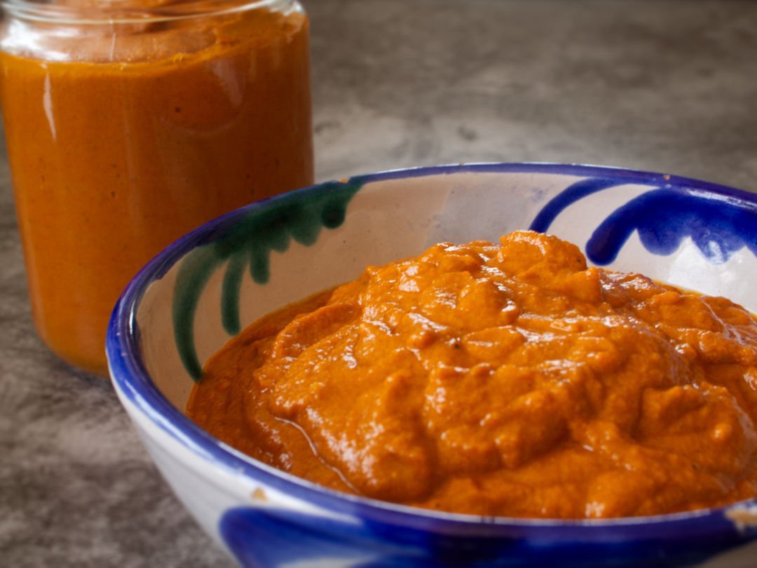 A small bowl of romesco sauce sits with a jar full of romesco sauce in the background