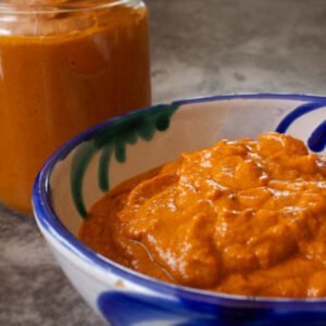 A small bowl of romesco sauce sits with a jar full of romesco sauce in the background