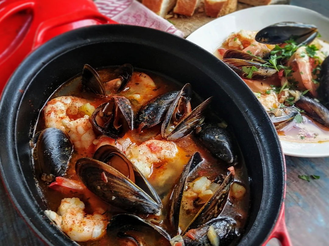 A large dish of seafood stew sits on a table beside a bowl of stew