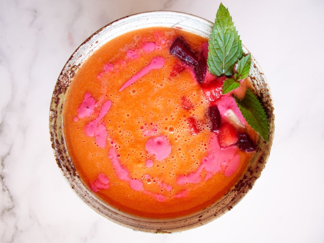 A small ceramic bowl sits filled with water melon gazpacho that has been garnished with a few sprigs of mint