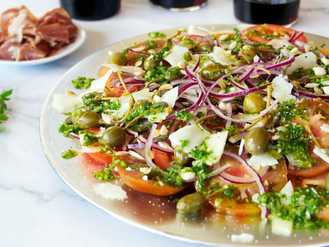 A large plate of Mediterranean Tomato Salad sits. with lots of pesto dressing and shaved pieces of Manchego cheese