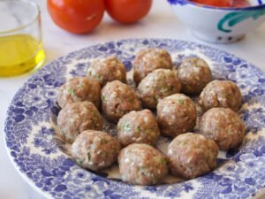 Uncooked albondigas sit on a pattered blue plate beside some tomatoes and extra virgin olive oil