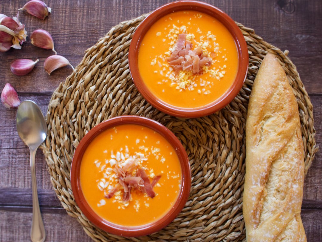 two bowls of salmorejo sit beside a loaf of bread and a few cloves of garlic