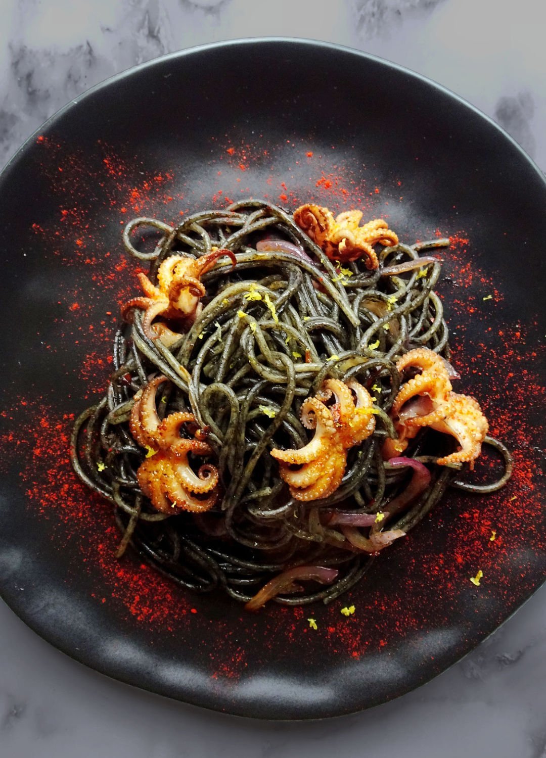 a black plate sits on a granite bechtop filled with black squid ink pasta and garnished with red baby octopus.