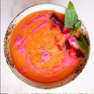 A small ornamental bowl is filled with a bright pink watermelon gazpacho and garnished with some sprigs of fresh mint