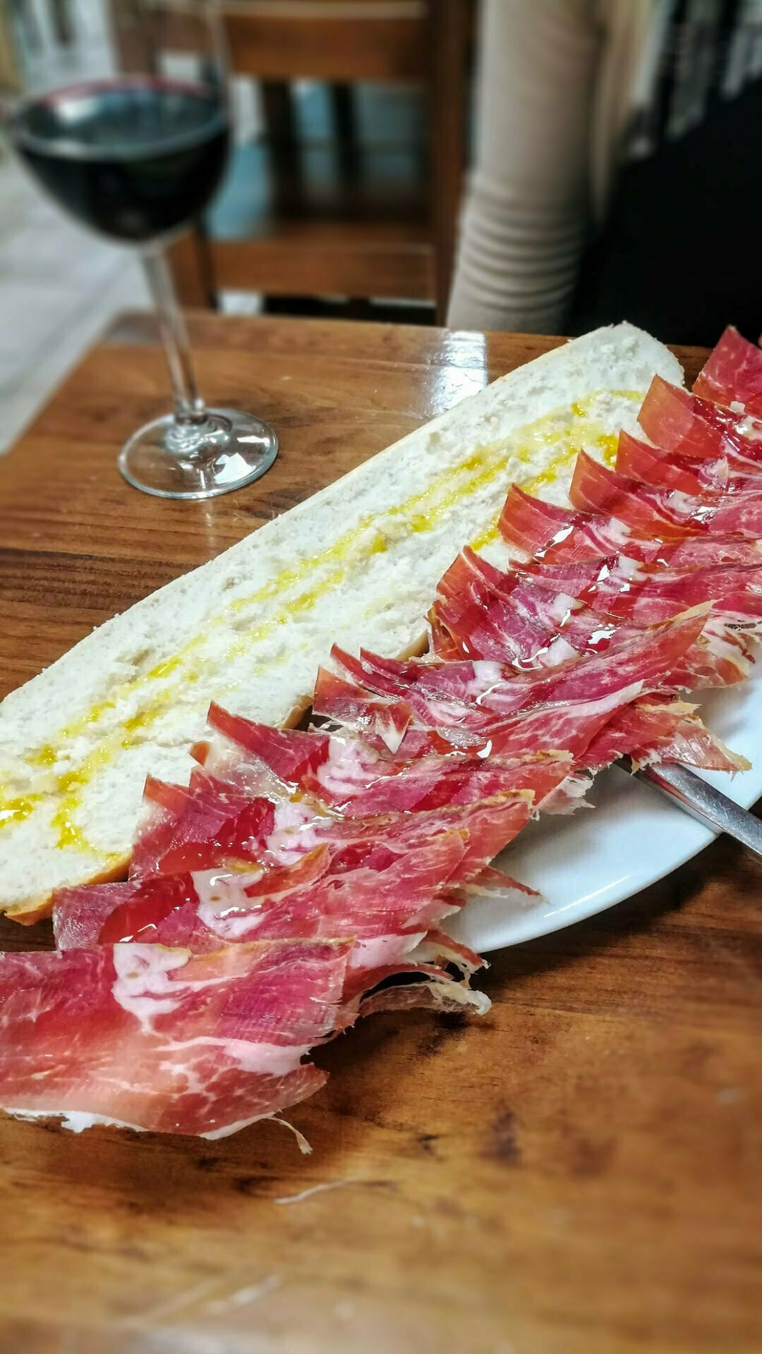 a large bocadillo with slices of ham sits on a plate next to a glass of red wine. 