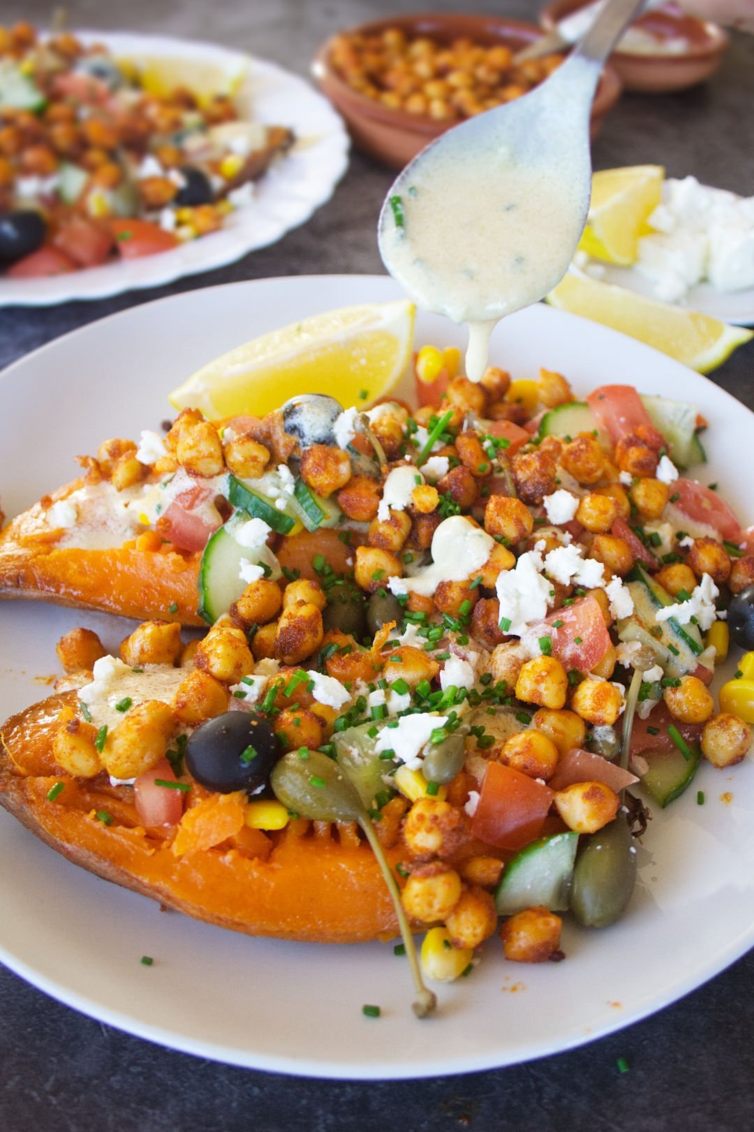 a large baked sweet potato sits on a white plate with plenty of Mediterranean inspired toppings.