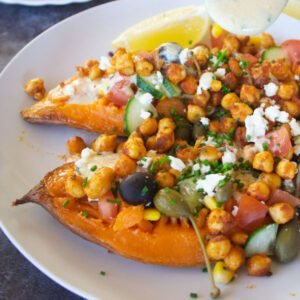 a large baked sweet potato sits on a white plate with plenty of Mediterranean inspired toppings.